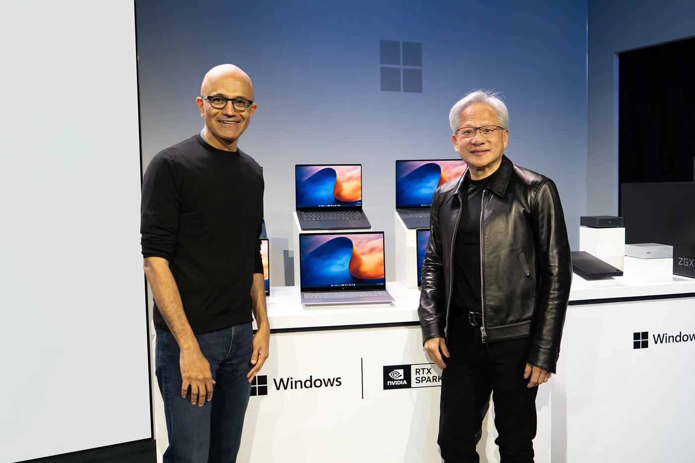 Satya Nadella and Jensen Huang on a stage in front of a wall of RTX Spark laptops and a Windows sign, with DGX Station boxes on the right.